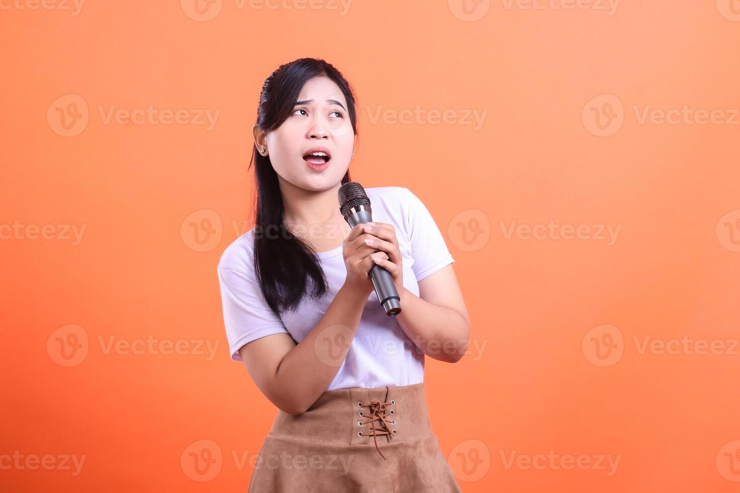A young Asian woman singing with a microphone, looking up with her mouth open in a performance pose, isolated on orange background photo