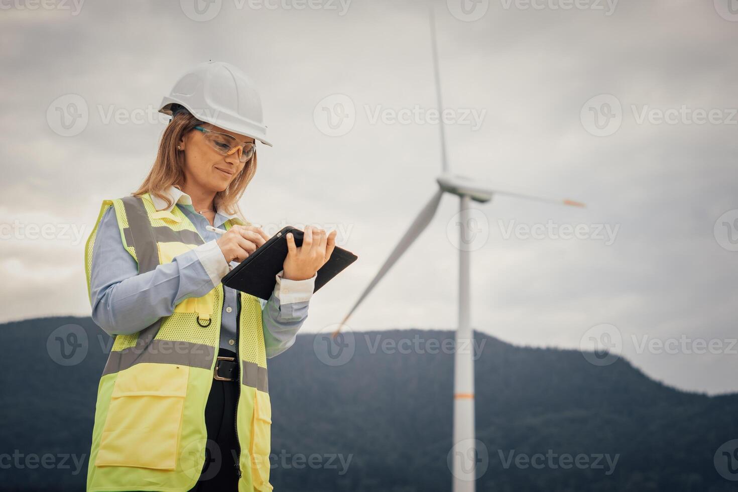 A female engineer in a safety helmet and yellow vest inspects data on a tablet at a wind energy site, surrounded by tall wind turbines representing advancements in renewable energy photo