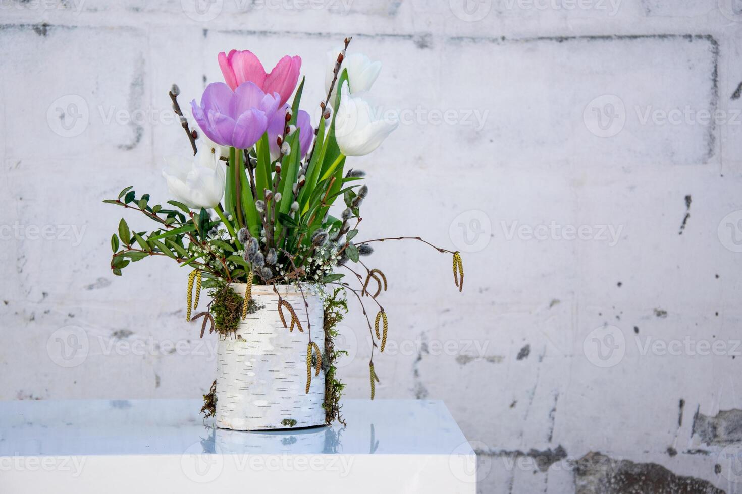 A vibrant arrangement of tulips in shades of pink and white features greenery and moss in a decorative container near a textured wall photo