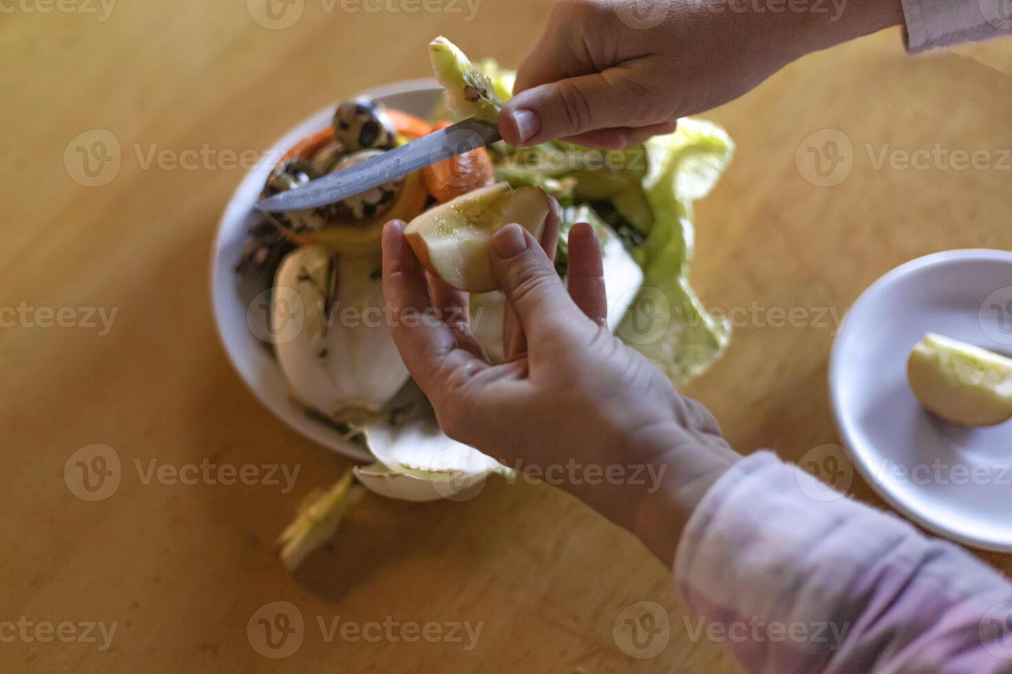 hands peeling an apple with a knife over a food waste container photo
