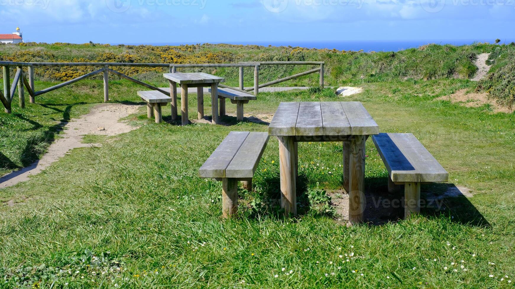 Wooden picnic tables and benches are set in a grassy open field under a bright blue sky, with a rustic wooden fence and a scenic coastal view in the background photo