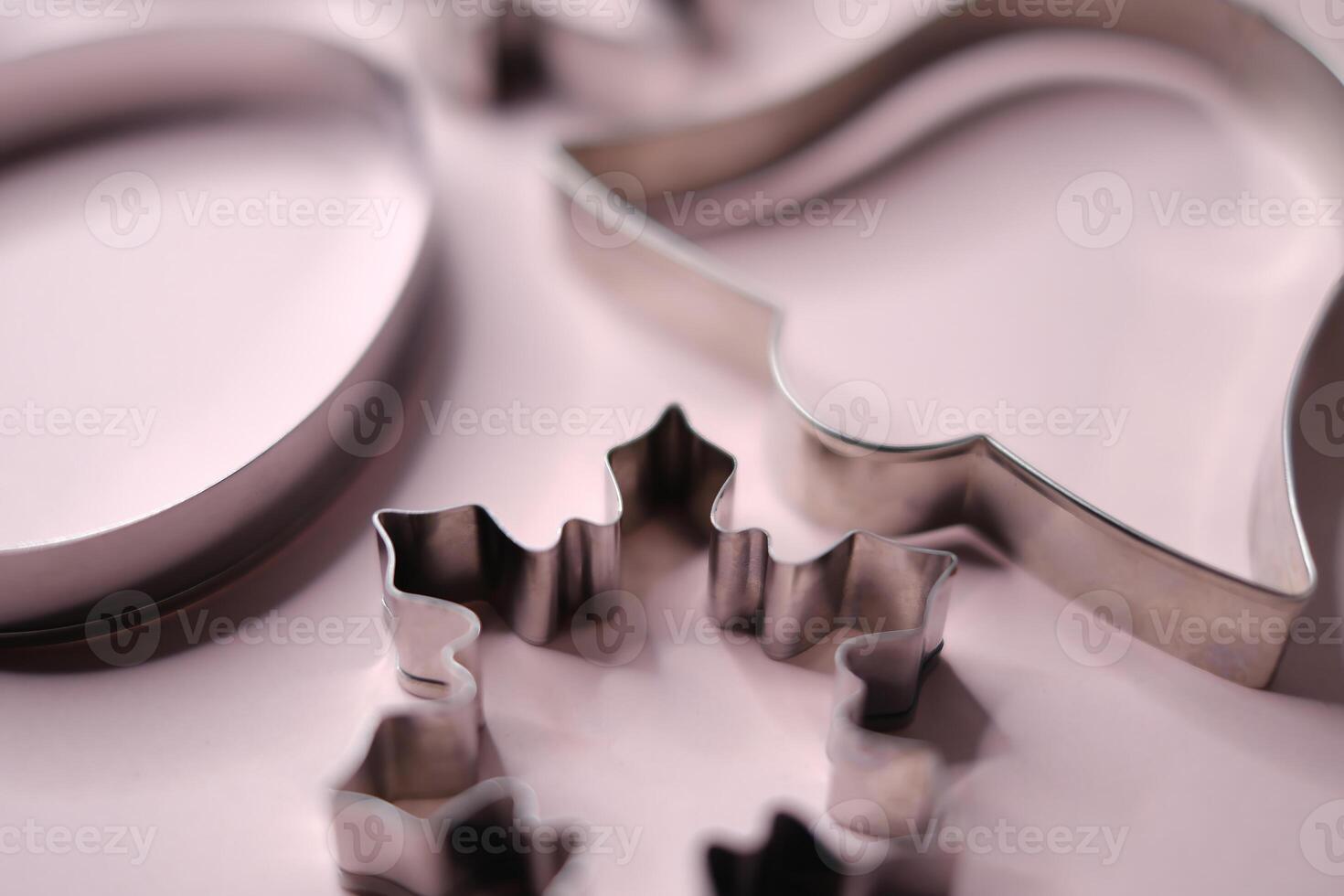 Close-up view of stainless steel cookie cutters in festive shapes including a snowflake, bell, and circle, placed on a soft pink background. photo