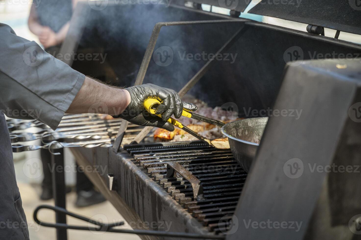 Male chef preparing meat on grill at barbecue party in summer garden, chef's hands in gloves. Concept of food, people and family time. Pork on skewers is roasted on coals in large metal grill device photo
