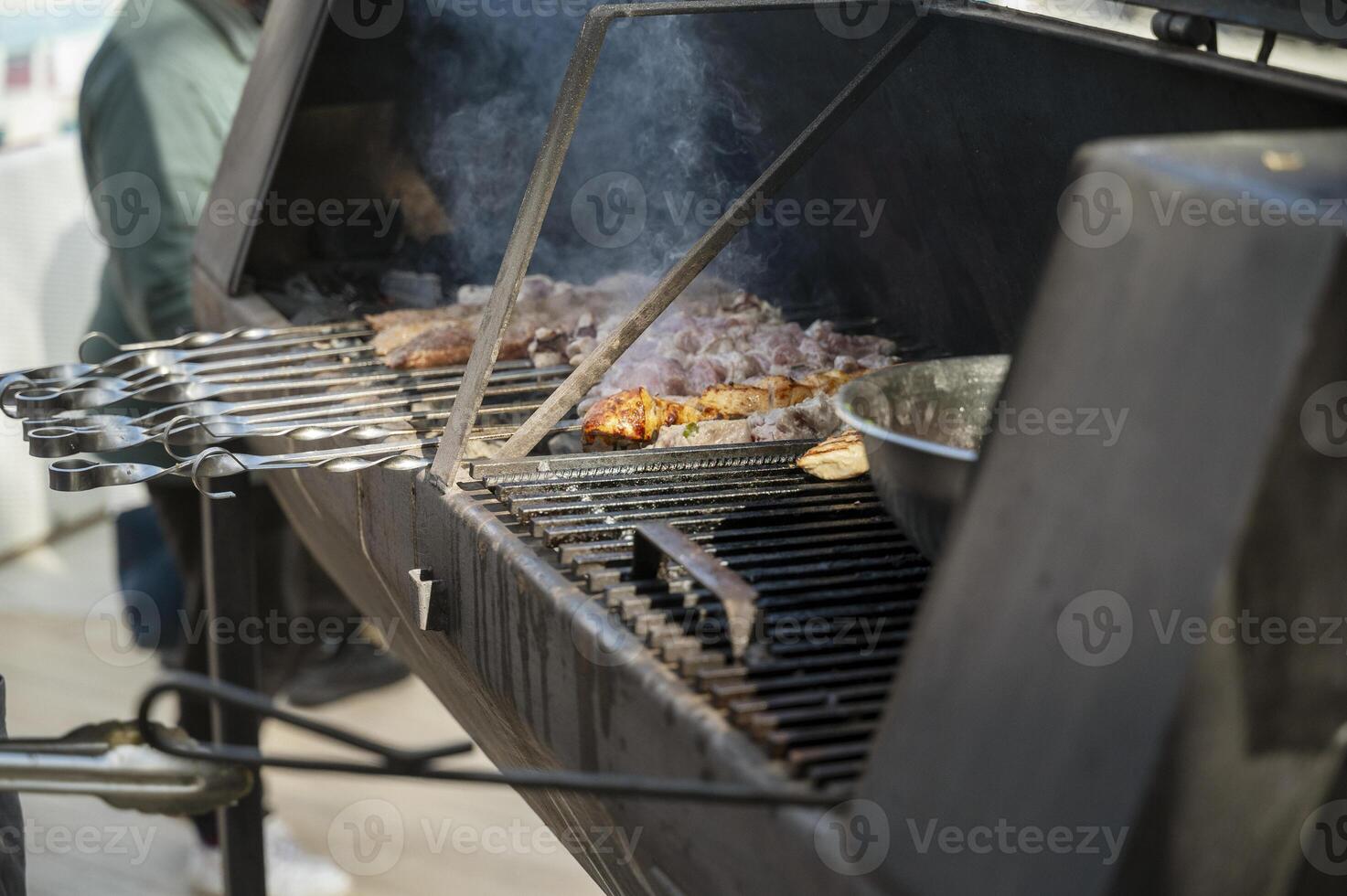 cooking meat on grill at barbecue party in summer garden. Food, people and family time concept. pork on skewers roasting on coals in large metal grill device. smoke from fire photo