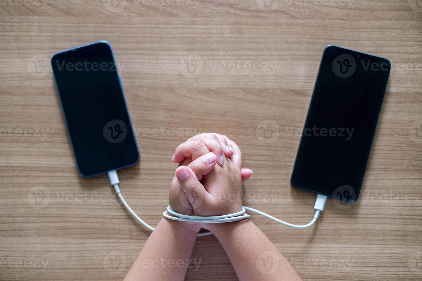 Child's Hands tied with charging cables between two smartphones on wooden table, symbolizing smartphone addiction and dependency in modern digital life. Conceptual image for technology overuse photo