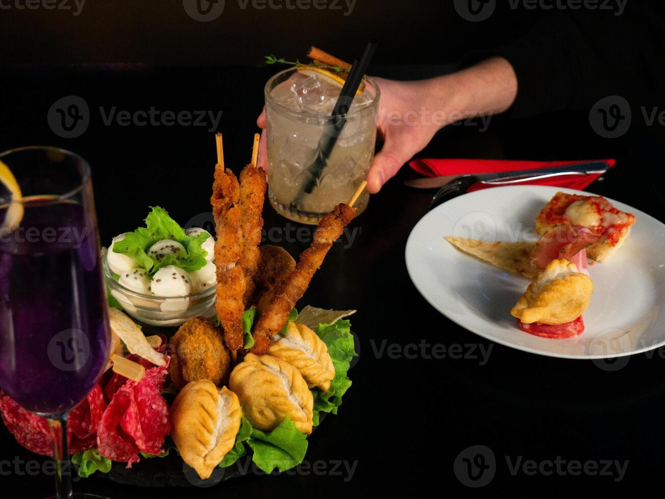 Close-up of a table with two cocktails, a glass of purple drink and a clear mixed drink held by a hand, and plates of various appetizers including skewers and mozzarella. photo