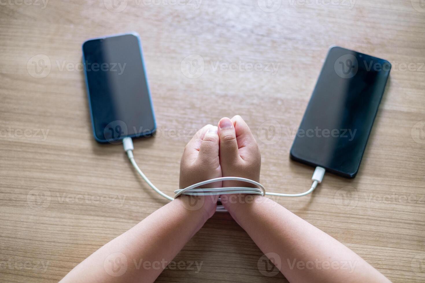 Child's Hands tied with charging cables between two smartphones on wooden table, symbolizing smartphone addiction and dependency in modern digital life. Conceptual image for technology photo