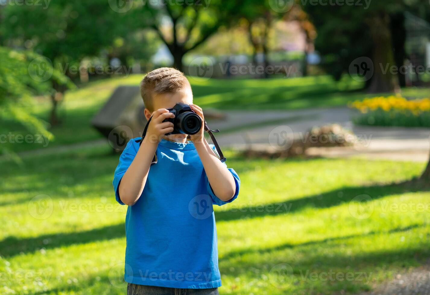 View of cute boy in blue t-shirt taking pictures with camera in city park photo
