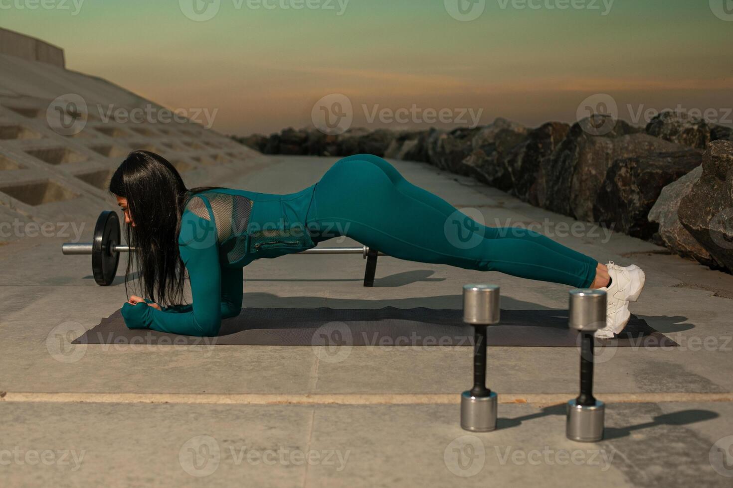 person engages in plank exercise on fitness mat near shoreline during early morning. tranquil waters and rocks create serene atmosphere for workout photo