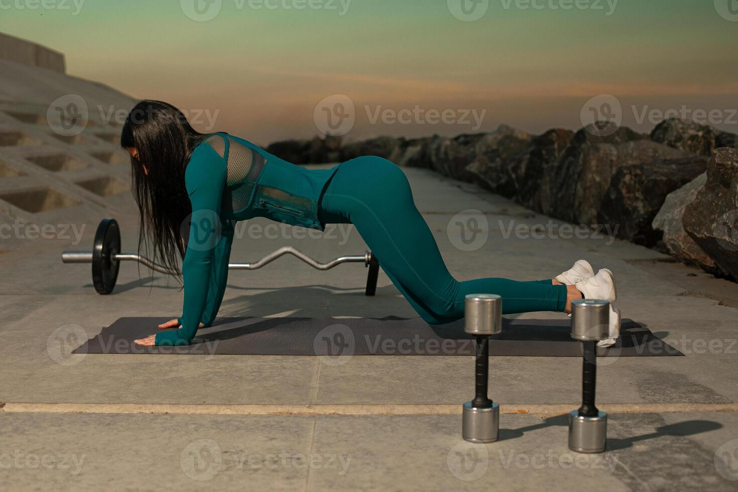 fitness enthusiast dressed in teal activewear performs push up exercises on mat positioned on rocky shoreline. Dumbbells are placed nearby as sun sets creating serene backdrop photo