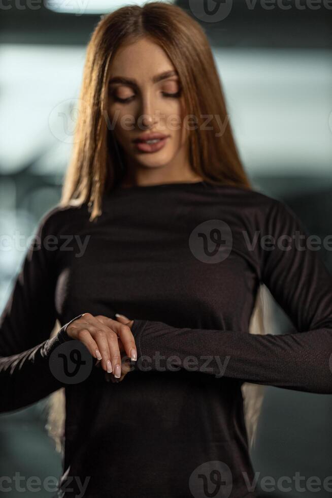 model stands in gym adjusting her long sleeve workout top while glancing her watch. environment is modern soft lighting creating athletic ambiance photo