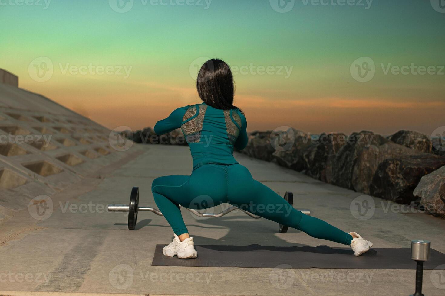 fitness enthusiast in teal athleisure performs squat on mat along rocky shoreline. sunset creates vibrant backdrop enhancing active atmosphere photo