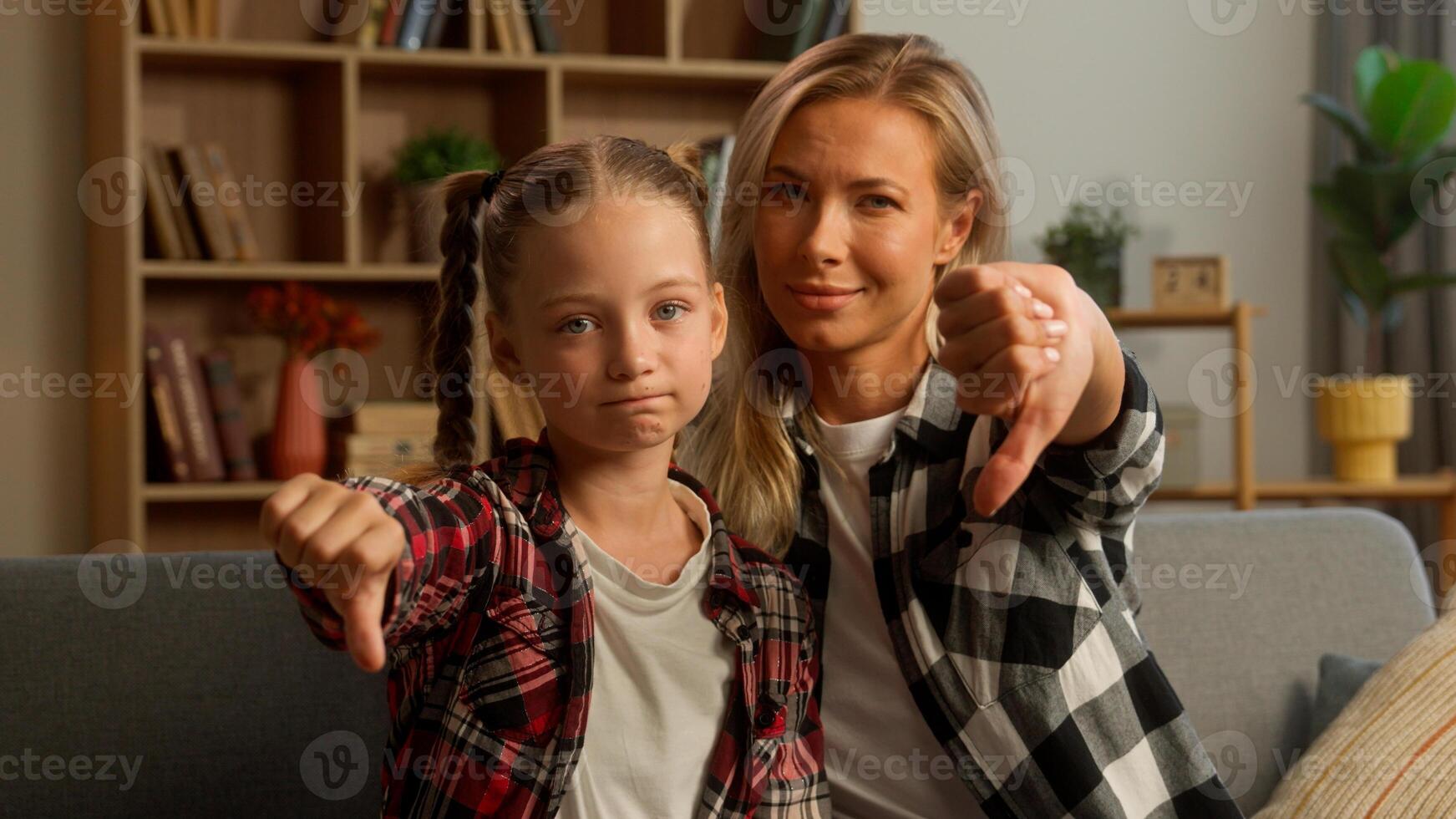 Caucasian family child schoolgirl looking at camera demonstrating thumb down upset little kid mother discontent sad mom woman female showing negative response parent daughter denial no gesture at home photo