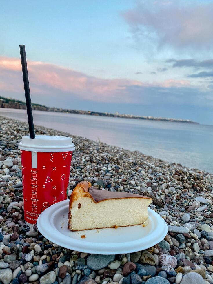 Cheesecake on a paper plate and takeaway coffee cup on a pebble beach with calm sea and pastel sky. Cozy outdoor snack scene in soft natural light. photo