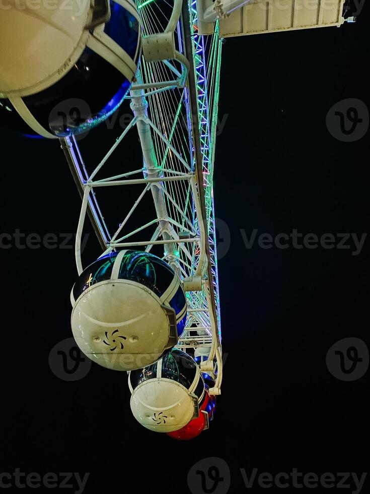 Low angle view of ferris wheel cabins illuminated with green, blue, and red neon lights at night against a dark sky, creating a vibrant and festive atmosphere perfect for travel and leisure themes photo