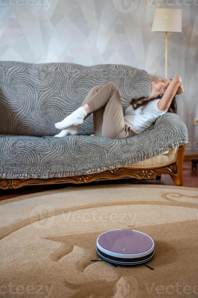 Robot vacuum cleaning the floor while a young woman relaxes on the couch in the background. Concept of smart home technology and effortless cleaning. High quality photo