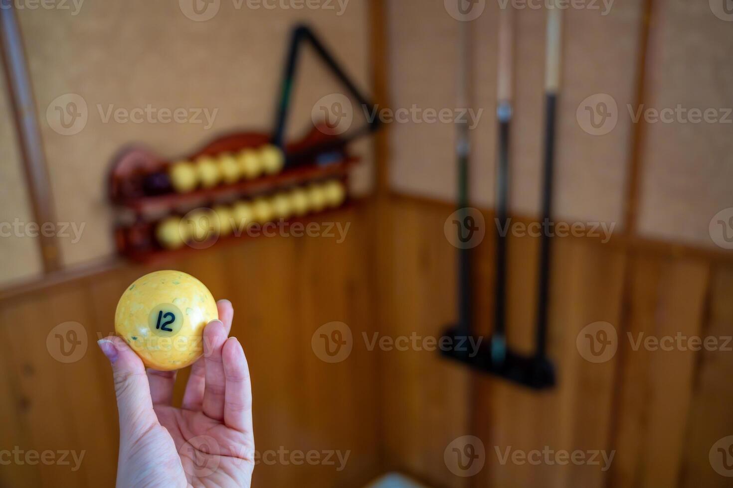 Hand holding billiard ball number 12 in front of a home billiard setup with cues and rack. Concept of indoor recreation and personal hobby space. High quality photo
