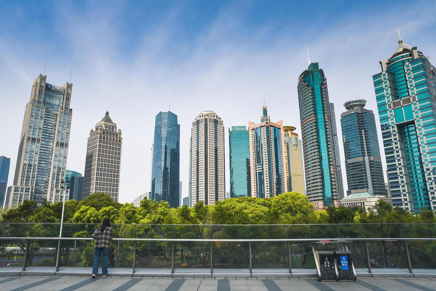 Shanghai, China - 1 April 2025 Modern Pudong business district with striking skyscrapers of diverse architecture against blue sky. A person admires the view from glass railings above lush urban park photo