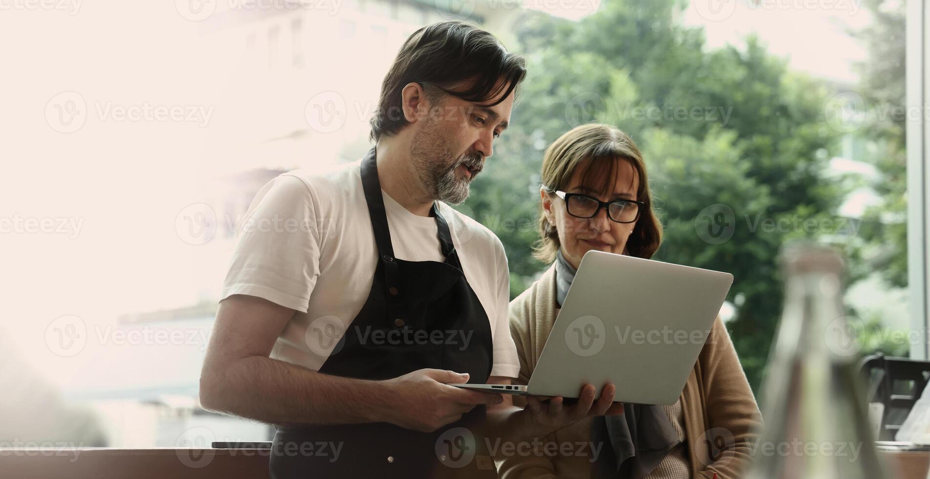 A friendly middle aged barista and a senior businesswoman collaborate using a smartphone and laptop inside a modern cafe. They appear to discuss online orders digital marketing or business strategy photo