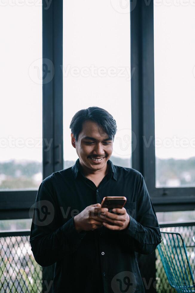 Smiling Asian man using smartphone near a window for communication photo
