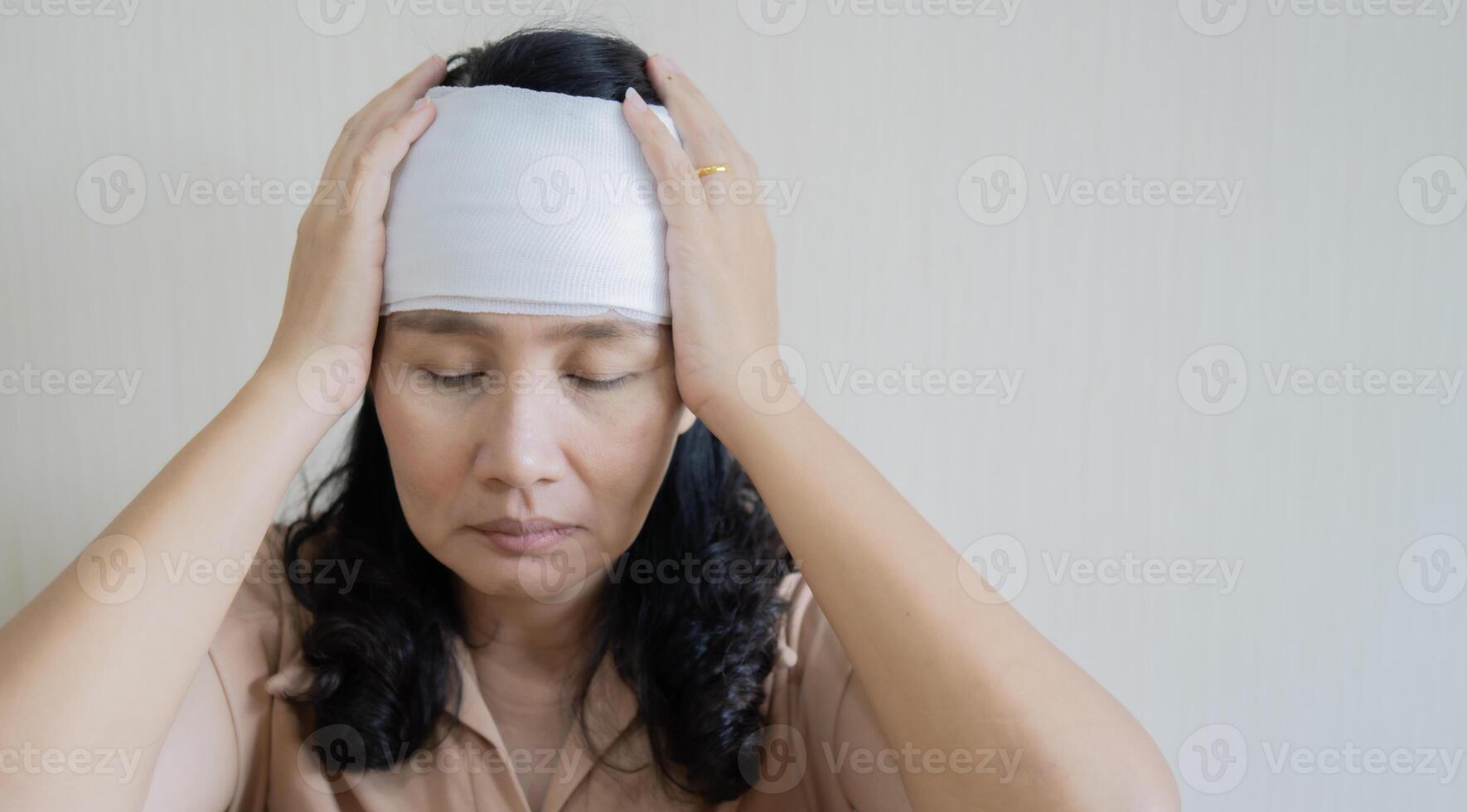 Portrait of a senior Asian woman close her eyes and touch her head with bandaged for an injury. photo