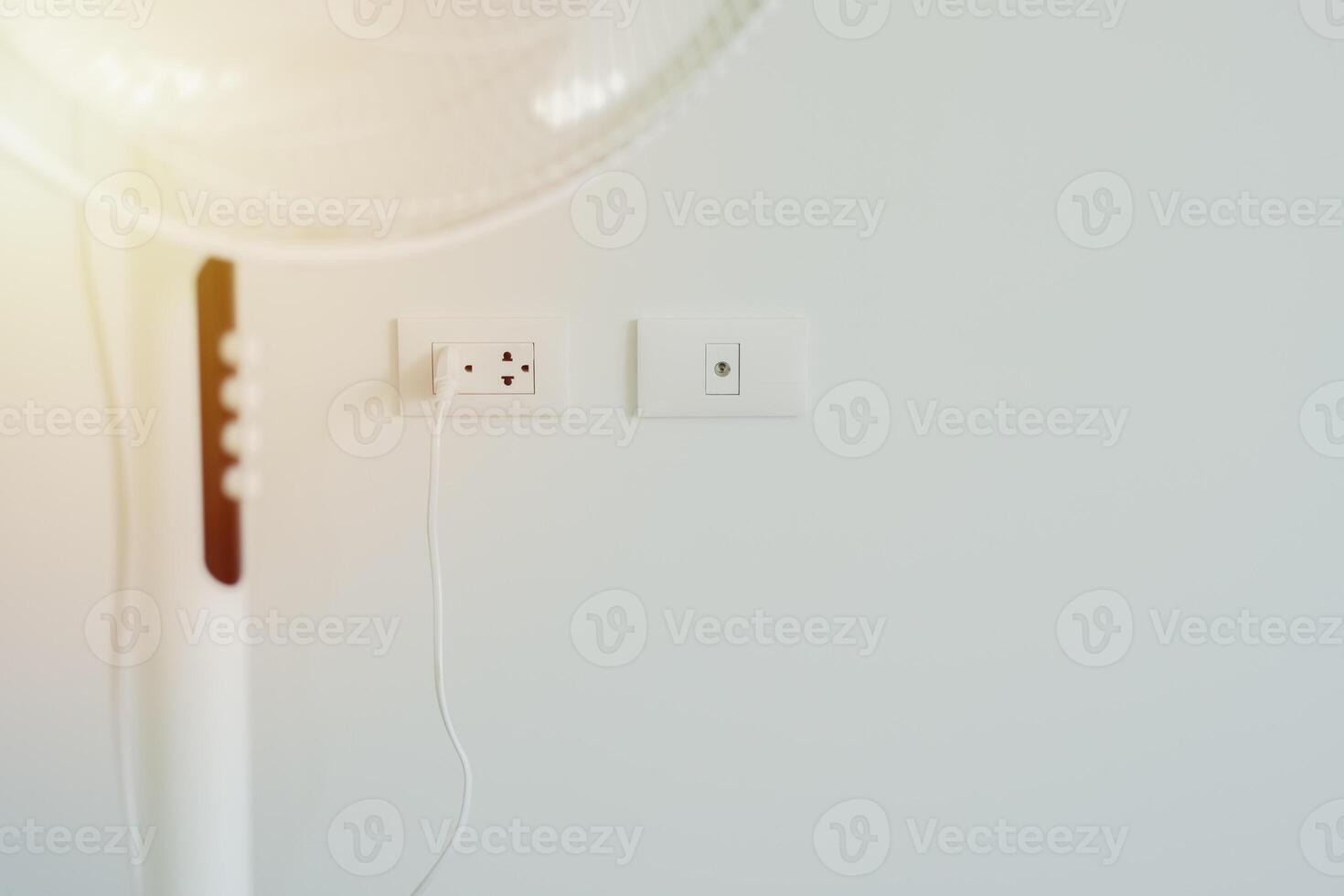 Cropped shot of white modern electric fan in light room with power plug on wall background. photo