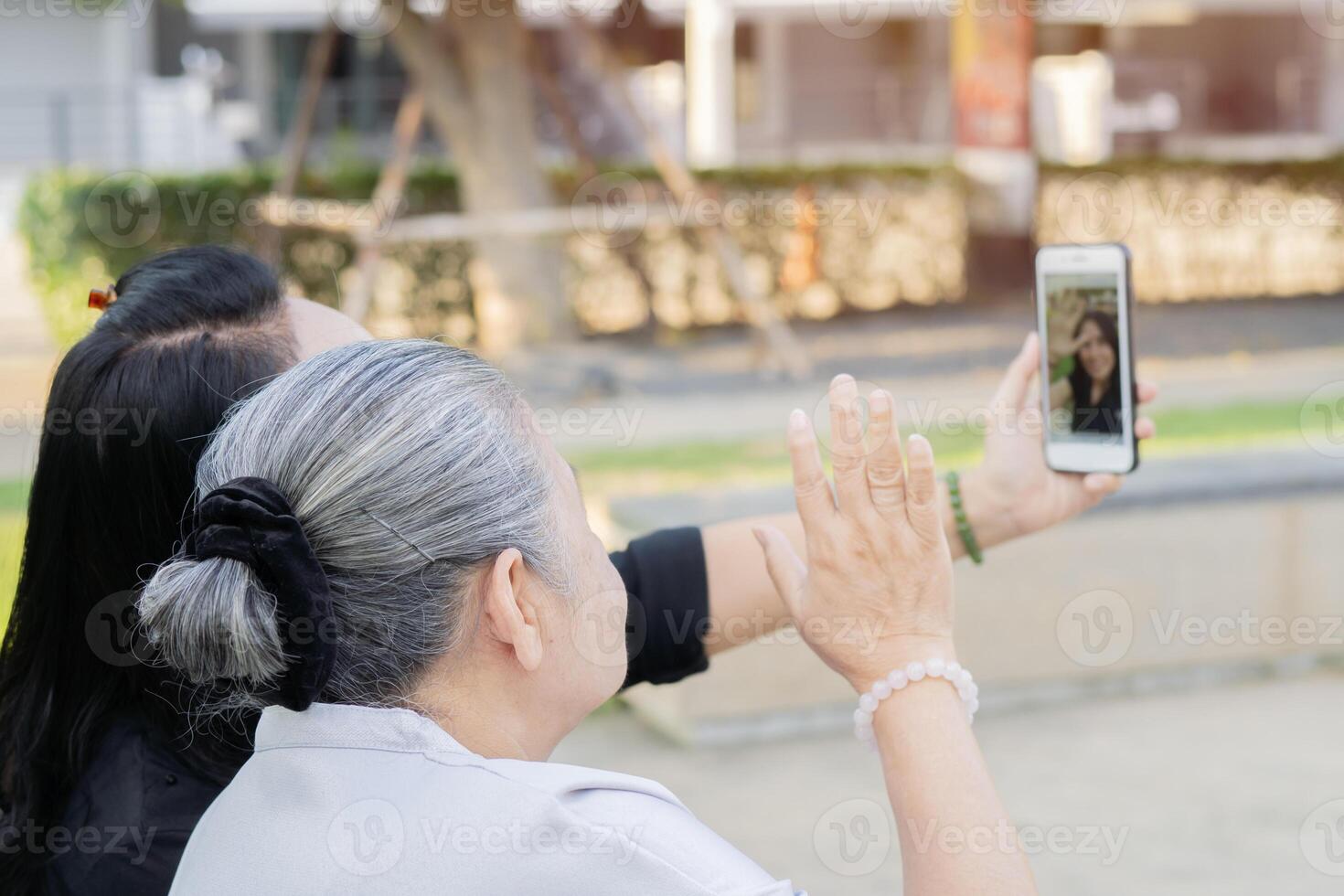 Happy Asian retired woman sitting on bench with daughter in park with using mobile phone photo