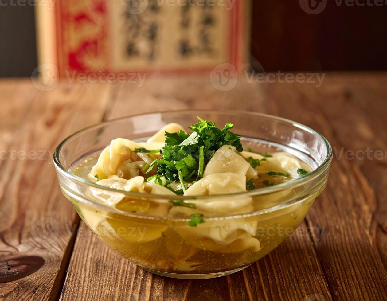 Wonton Soup with Dumplings, Herbs and Broth in a Clear Bowl on a Wooden Table photo