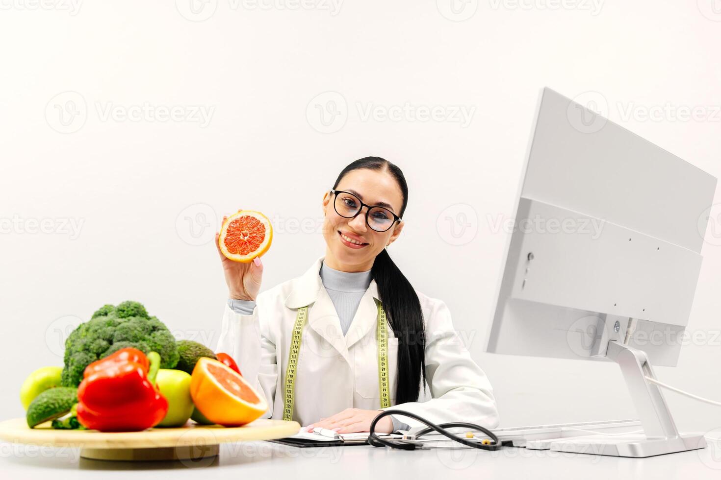A nutritionist in a lab coat with an avocado in hand sits at a table of fresh fruits and vegetables in a bright office of a modern clinic photo