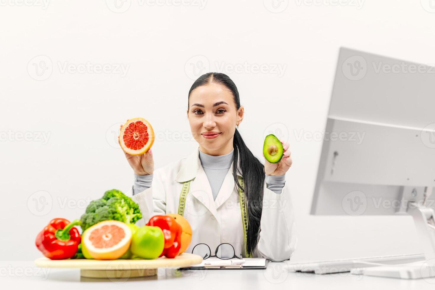 Young european woman dietician holding avocado and grapefruit, sitting at desk with fruits and vegetables, smiling at camera. Lady nutritionist recommending weight loss clinic, copy space photo