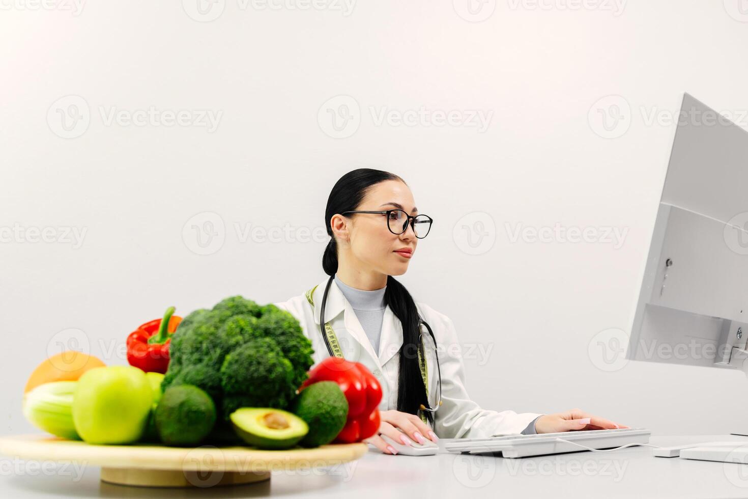 A nutritionist takes notes and works at a computer in a bright medical office. Fresh vegetables and fruits are on the table photo