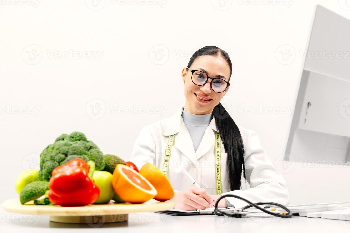 A young female nutritionist points out fresh fruits and vegetables. Broccoli, apple, avocado, celery, peppers, orange. Concept of the importance of healthy eating. White background photo