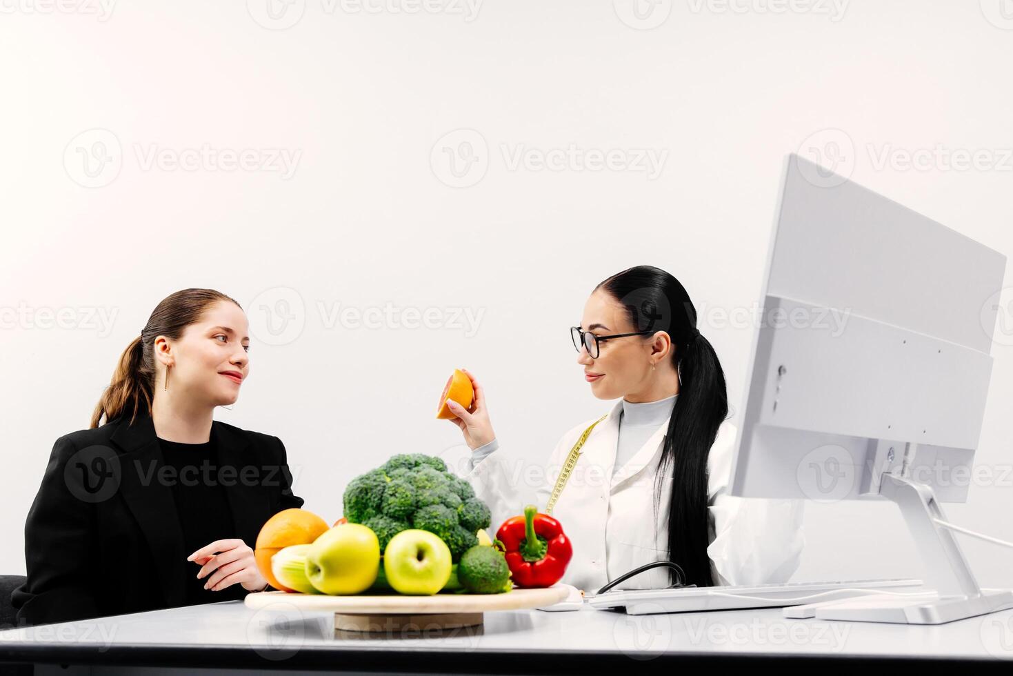 A nutritionist consults a patient, a young woman, in a modern clinic. On the table of the specialist doctor are fresh vegetables and fruits photo