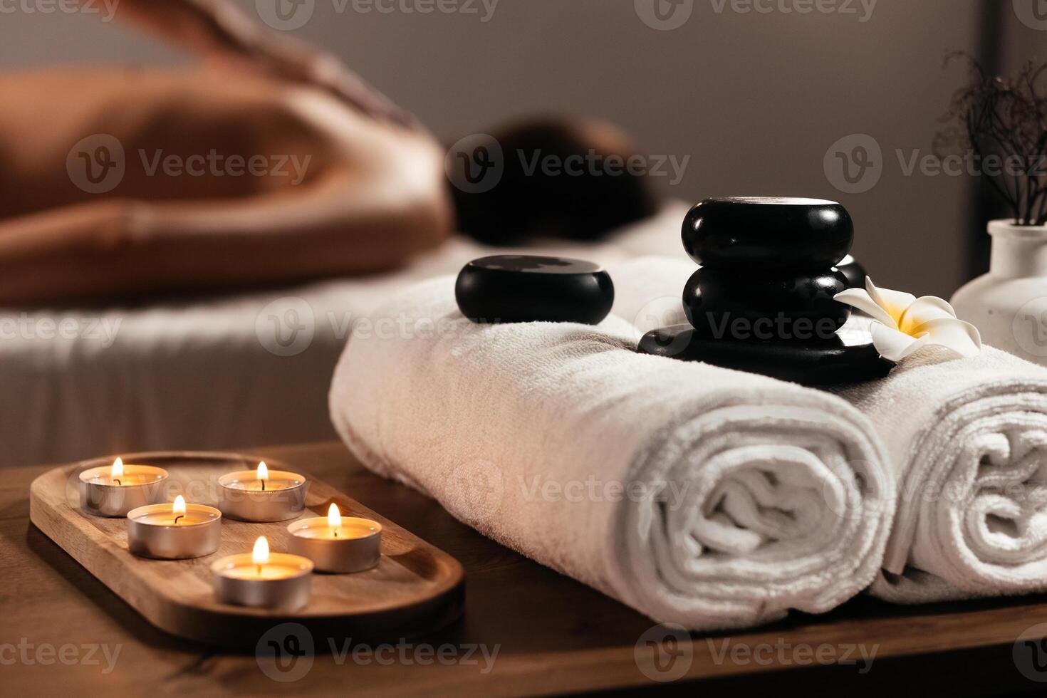 Woman enjoying exotic hot stones spa massage on blurred background. Spa supplies, burning candle and flower on table in beauty salon, space for text. Banner ads photo