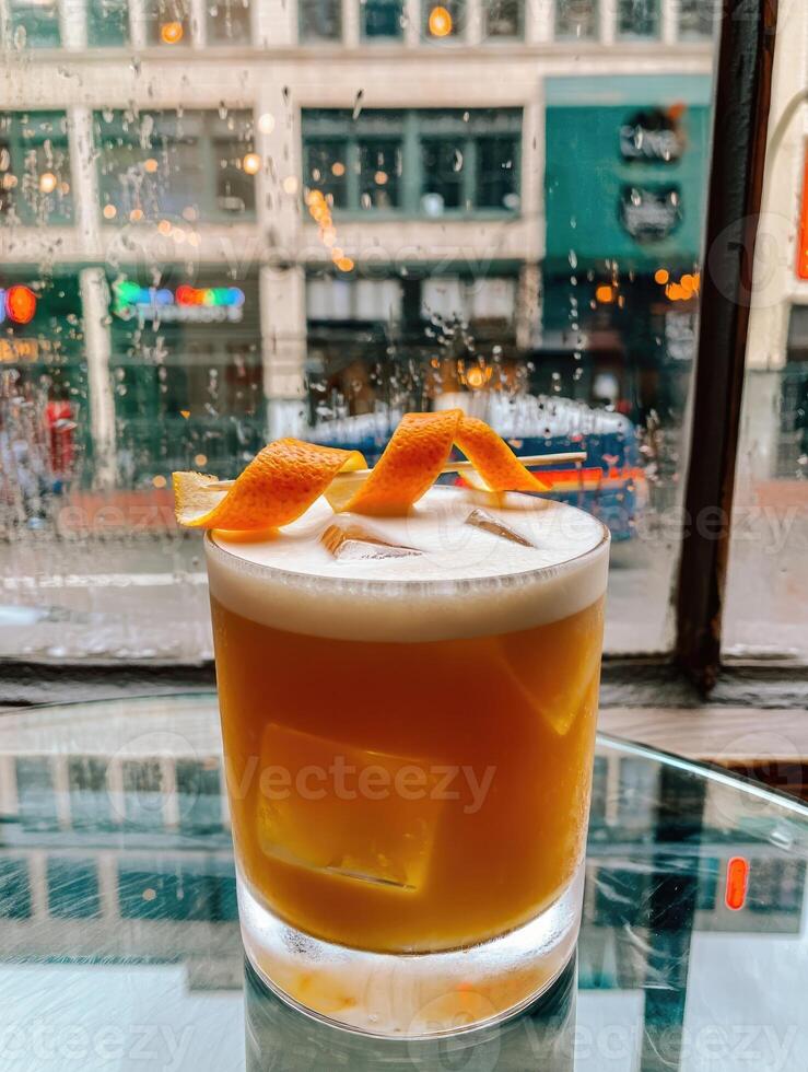 Craft cocktail with citrus garnish on a table near a rainy window in an urban environment photo