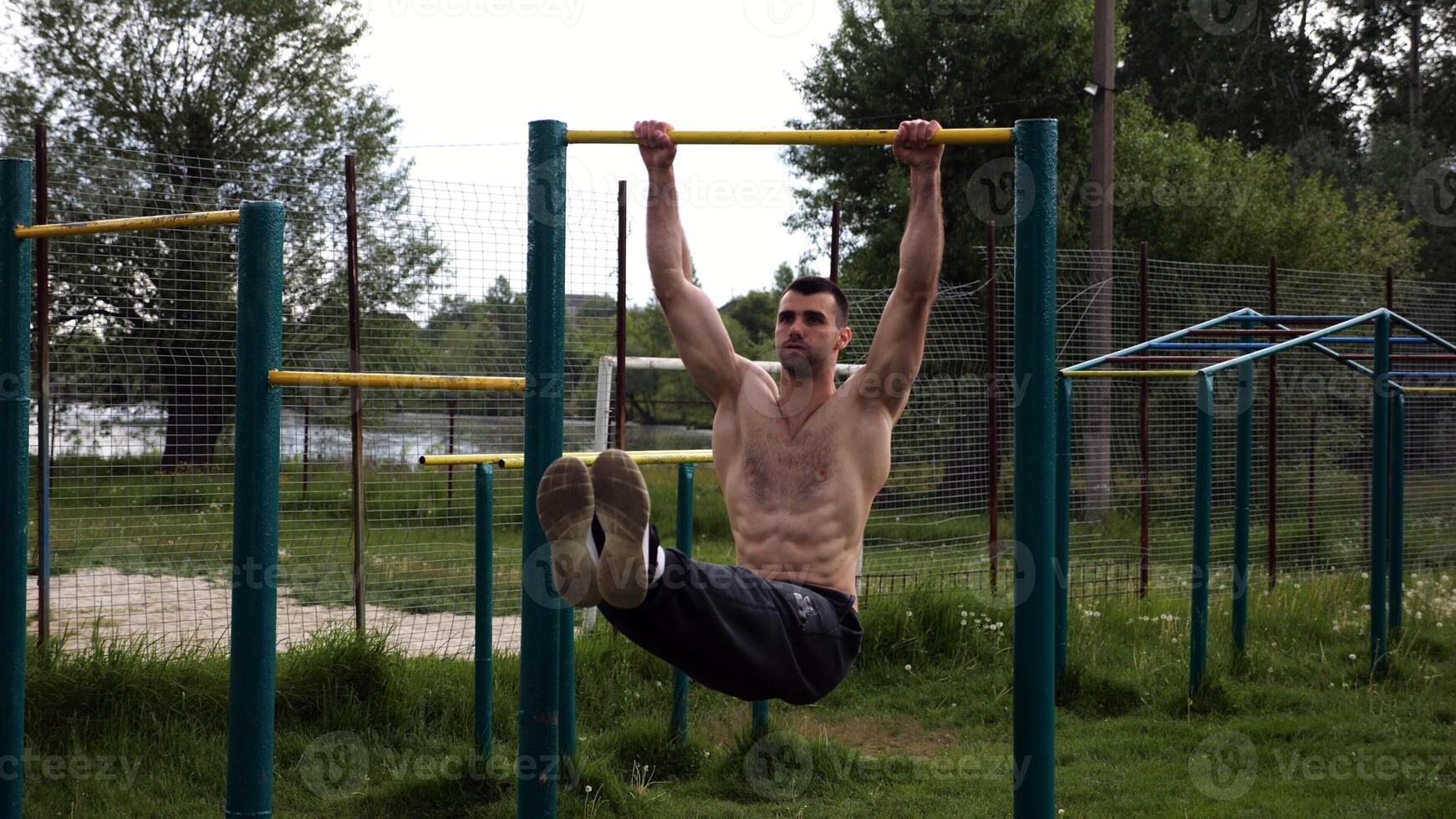 Athletic man trains abdominal muscles raising legs on horizontal bar at sports ground. Young sportsman exercising on pull-up bar outdoor. Sporty athlete working out at nature. photo