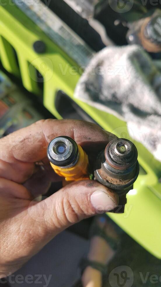 Hand Holding a New and an Old Fuel Injector in Front of Car photo