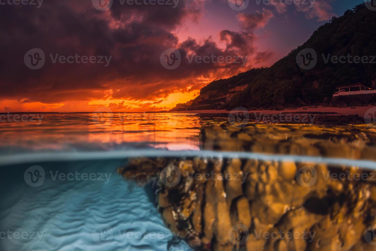 Dramatic sunset or sunrise over calm ocean with underwater sand patterns and coral, captured in a split view seascape photo