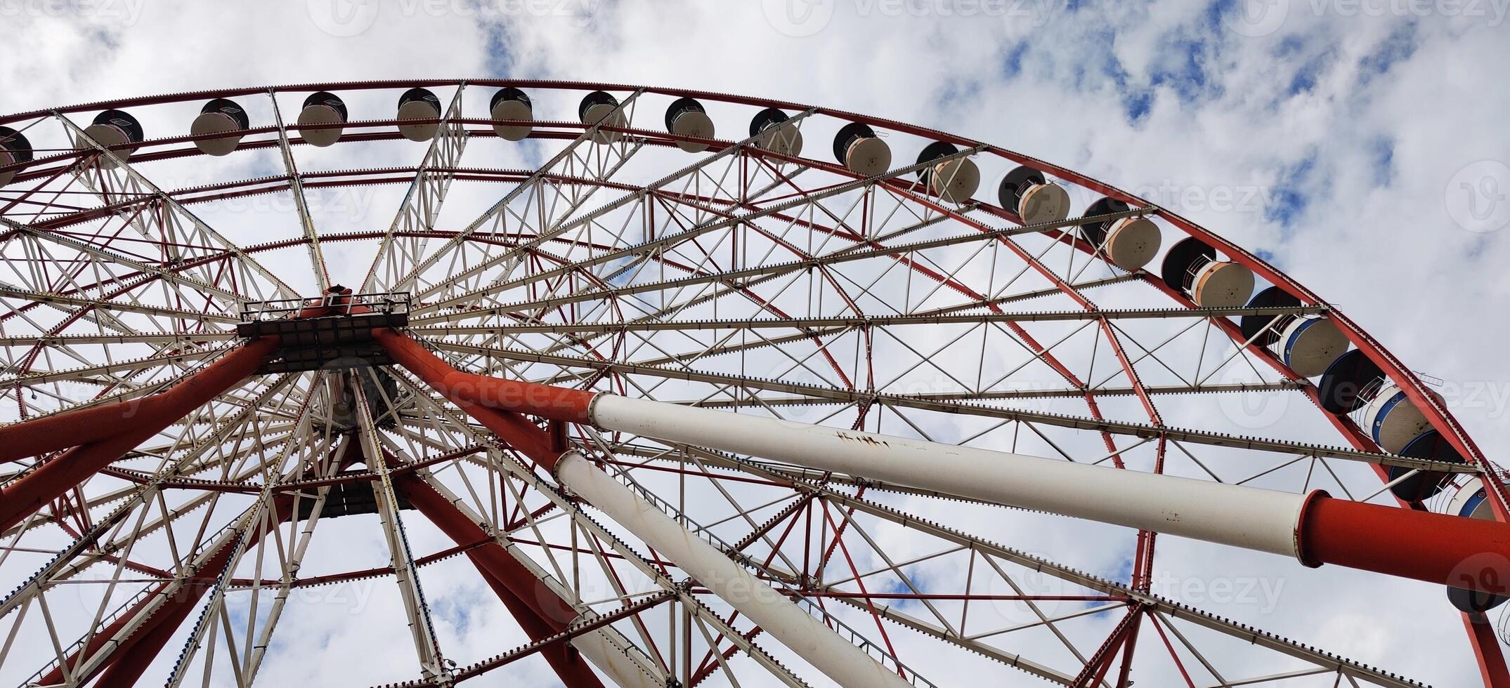 Low-angle view of a large Ferris wheel with red and white metal beams against a partly cloudy sky, showcasing its towering structure and passenger cabins. photo