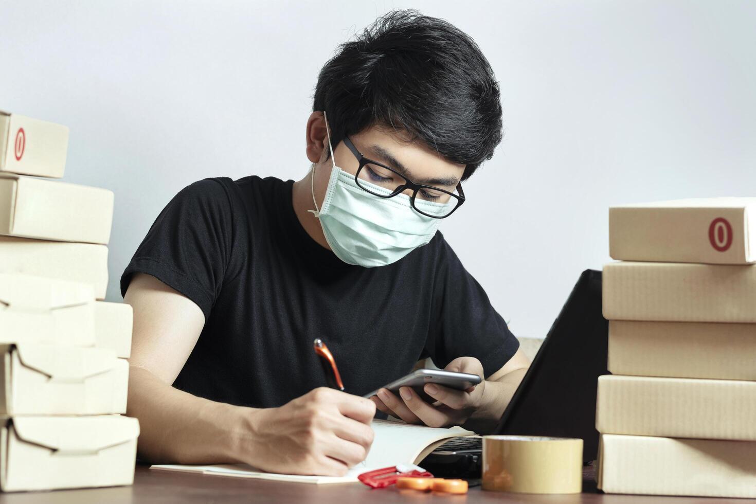 Asian man small business owner and Wear a mask to protect against Corona virus. Work from home, Have a laptop, and a smartphone recording the product information Online marketing, Startup SME photo