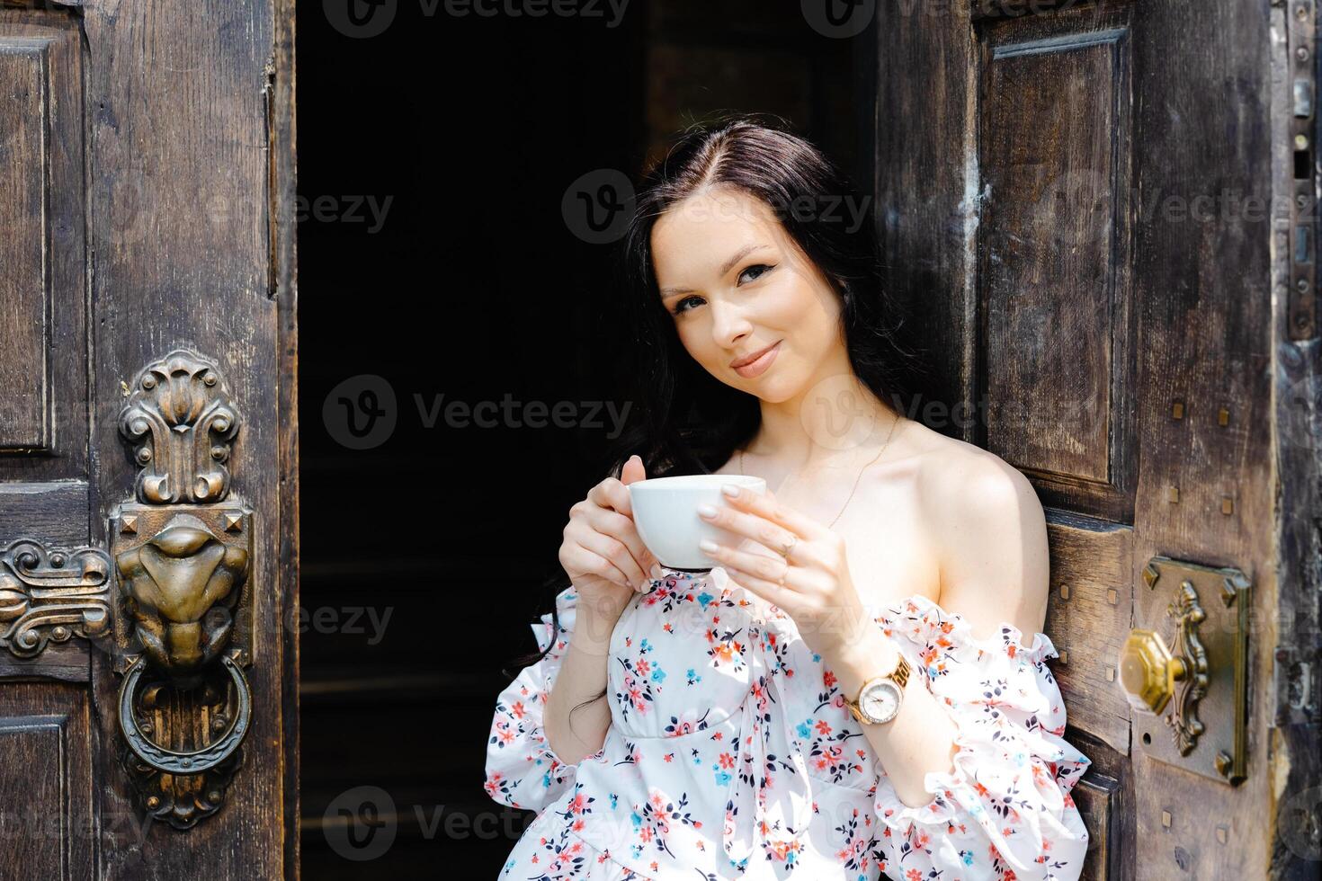 A young beautiful woman stands at the open old door of a coffee shop and holds a mug in her hands photo