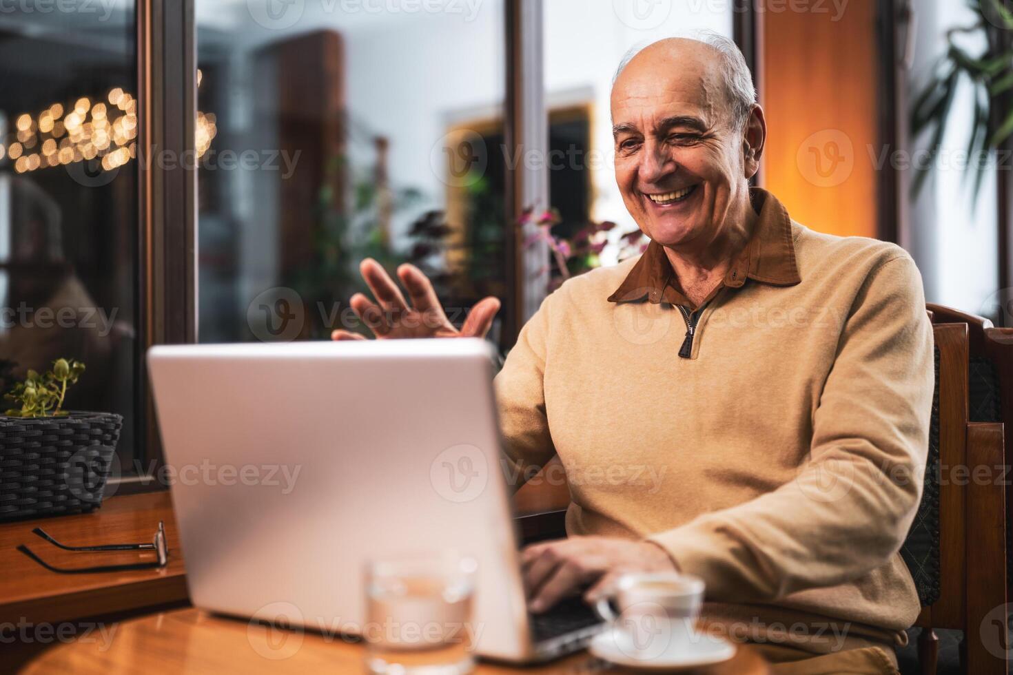 Happy senior male smiling and waving while talking during a virtual meeting on laptop in the bar. photo