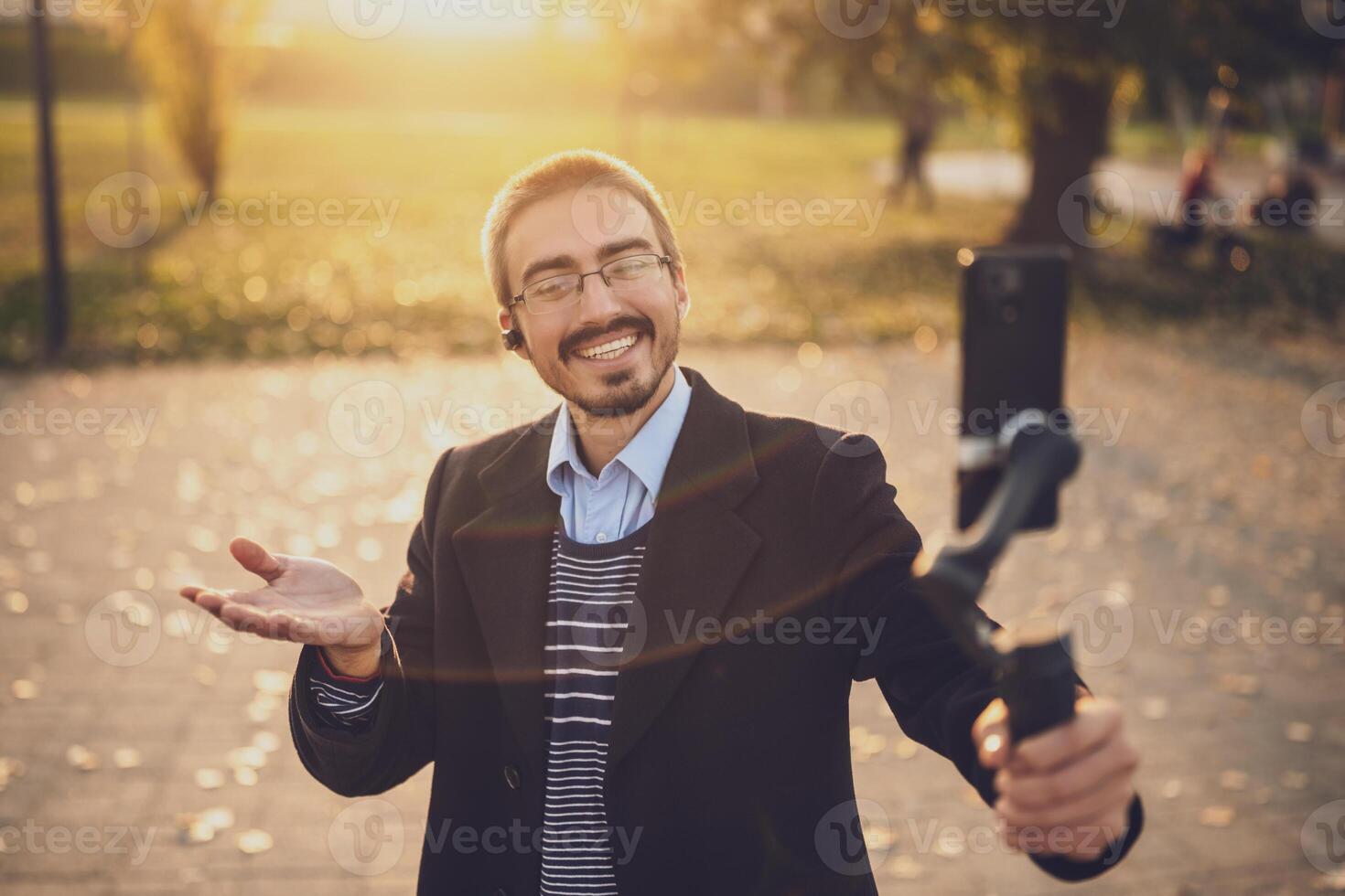 Male influencer explaining his business success through blogging, smiling and gesturing while recording with his smart phone and gimbal in a park. photo