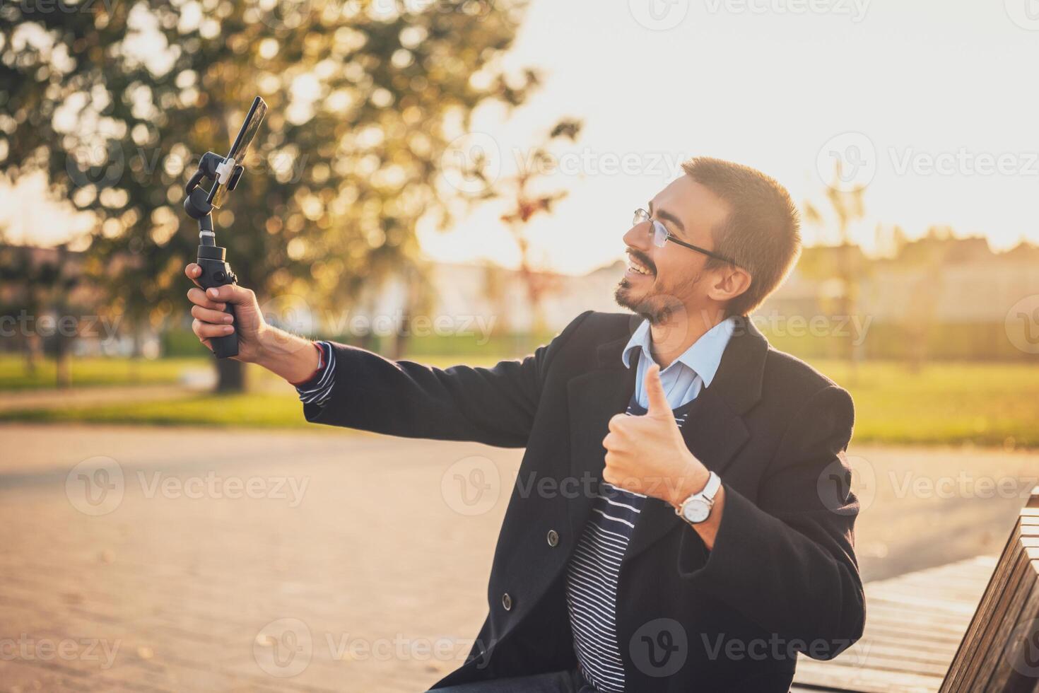 Confident male entrepreneur showing thumb up while recording business blog with gimbal stabilizer outdoor. photo