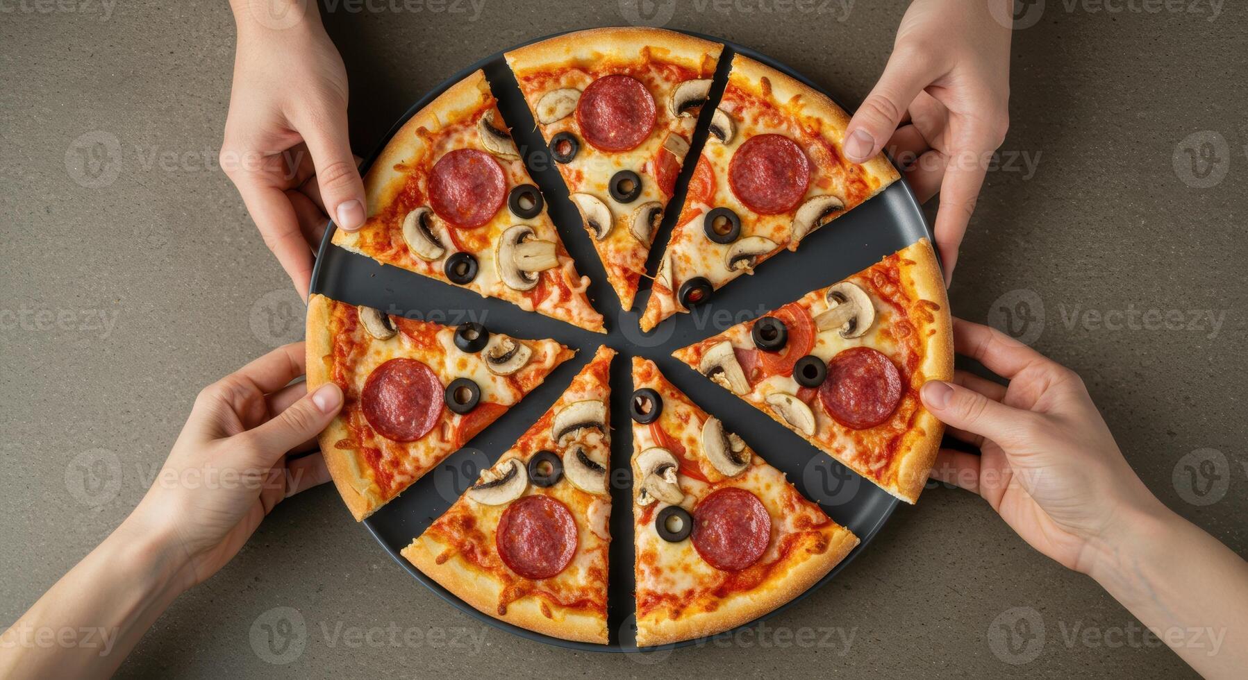 Top view of two pairs of hands holding a large round pizza cut into slices on a dark gray plate. Pizza is topped with pepperoni, mushrooms, and olives. photo