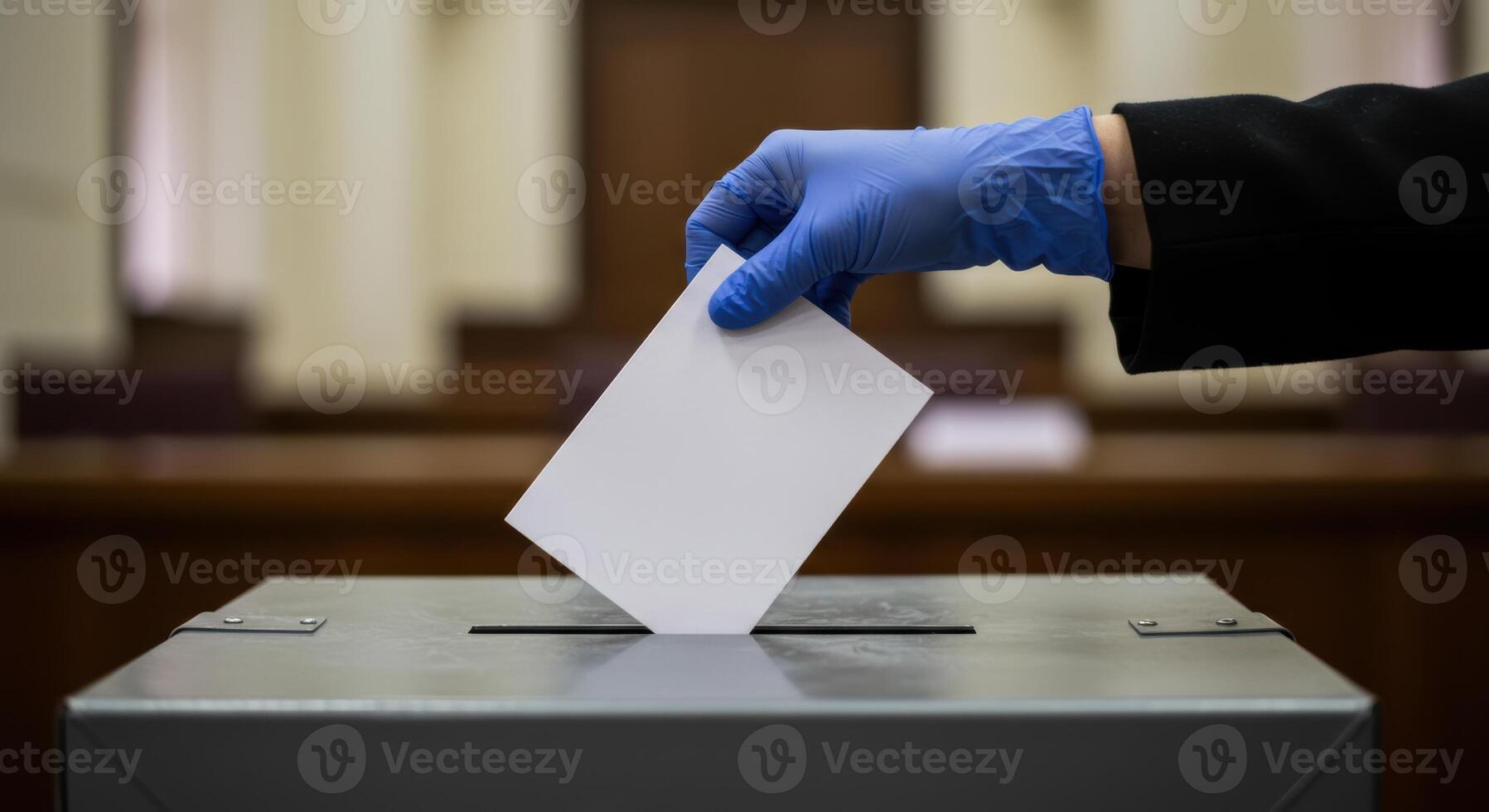 Gloved hand casts vote into ballot box during election. The ballot box is gray metal. Blurred background shows a civic building interior. photo