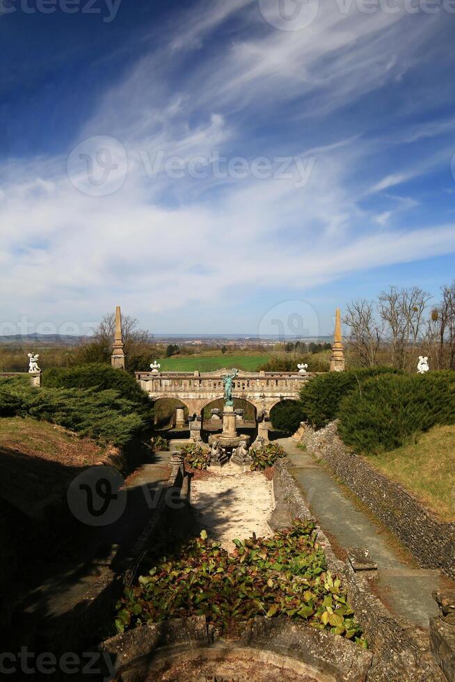 Ornate stone bridge at castle Roseburg framed by statues and green hedges with open countryside beyond photo