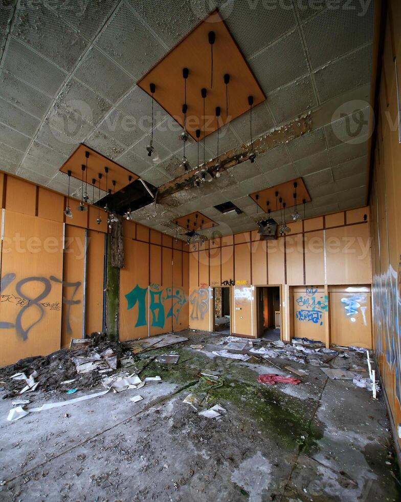 Decay and debris in an abandoned room with broken ceiling panels, graffiti, and scattered papers photo