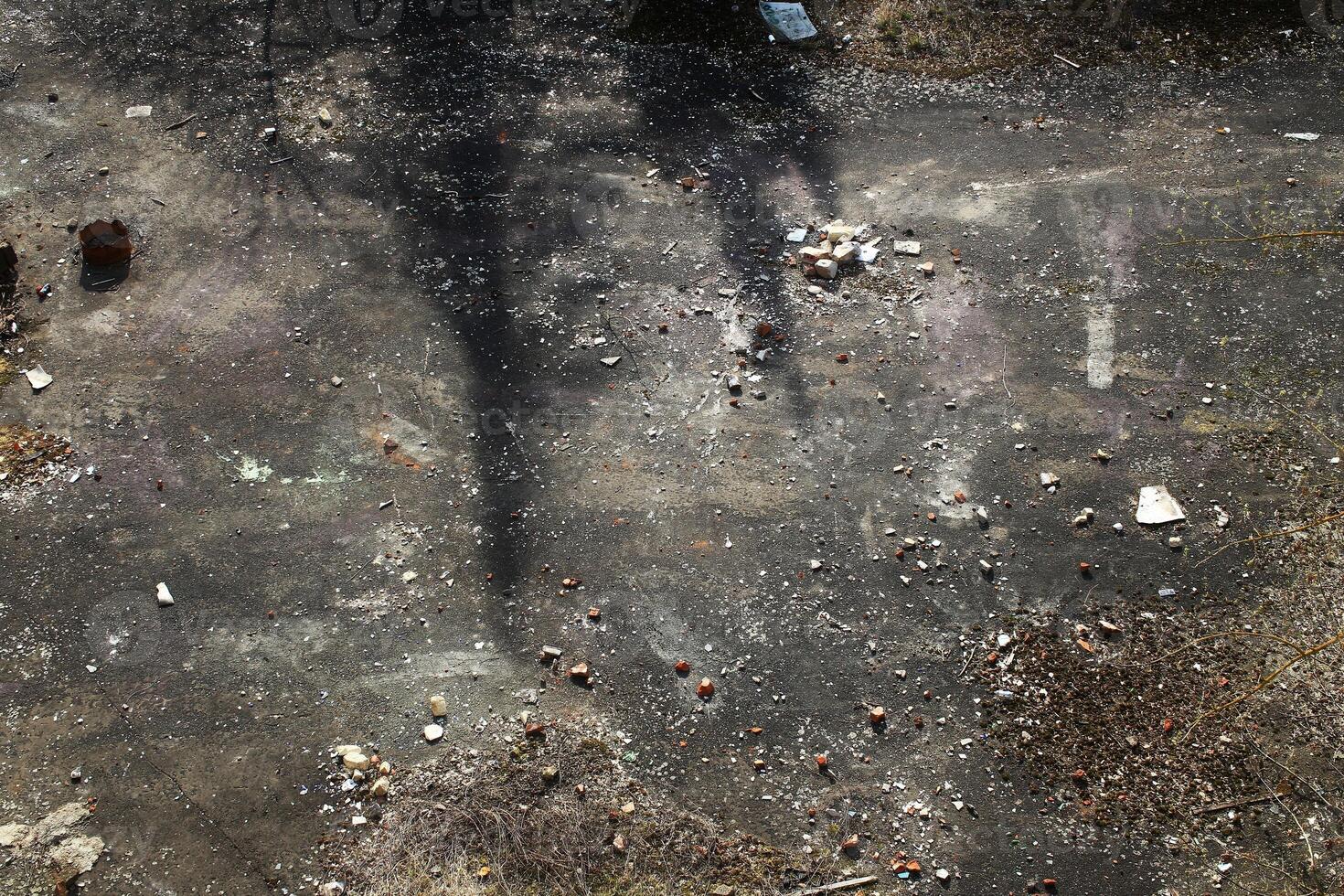Shadowed concrete ground with scattered rubble, dry brush, and faint surface markings photo