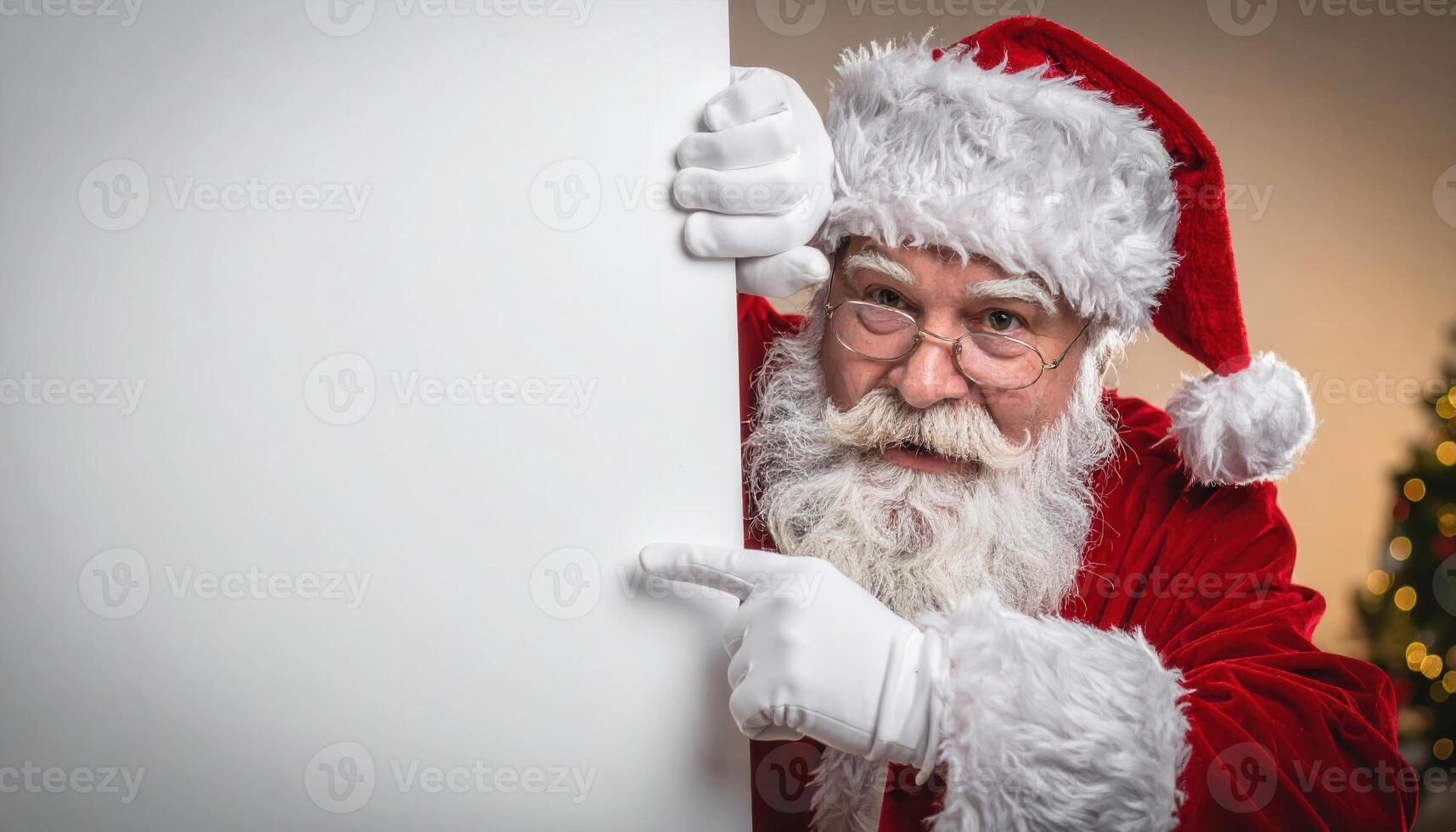 Santa Claus peeking from behind a large white sign, wearing red suit and hat, pointing to the blank space. Christmas tree in background. photo
